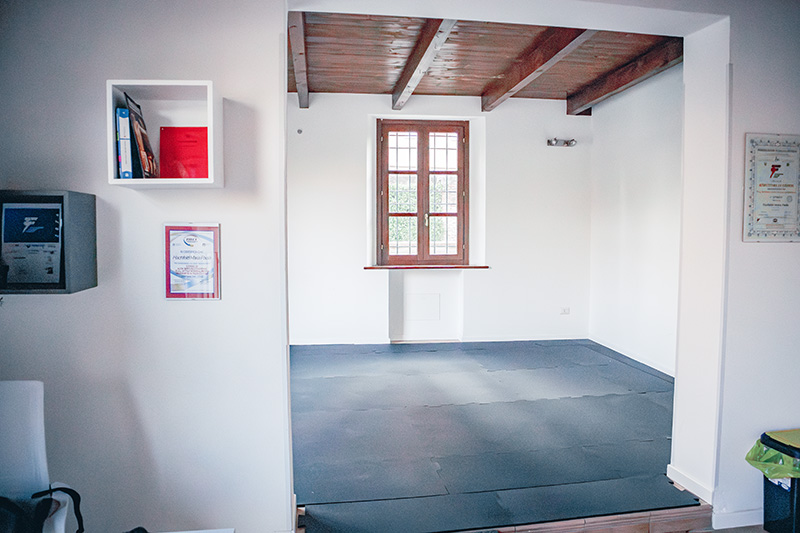 Sala per i corsi, gli allenamenti di gruppo e l'allenamento a corpo libero, con muri bianche, tetto in legno con travi a vista e pavimento di gomma grigio scuro.