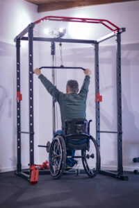 Atleta in sedia a rotelle di spalle che si allena nella rack in palestra, dimostrando l'accessibilità e l'inclusione nel fitness.